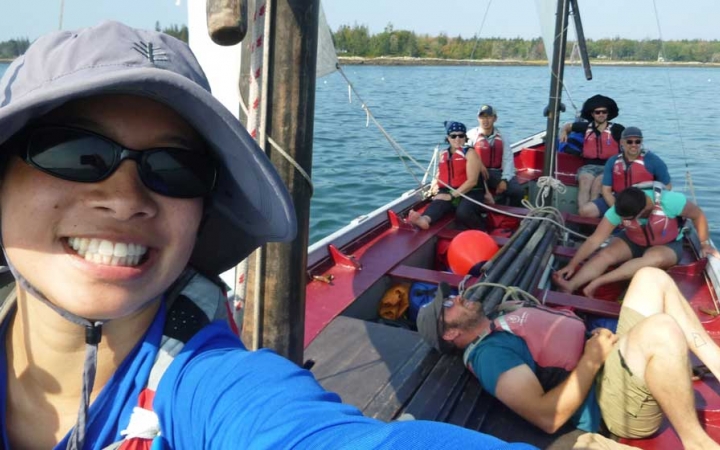 A young person takes a selfie while on a sailboat. Behind them, others are sitting in the boat. 